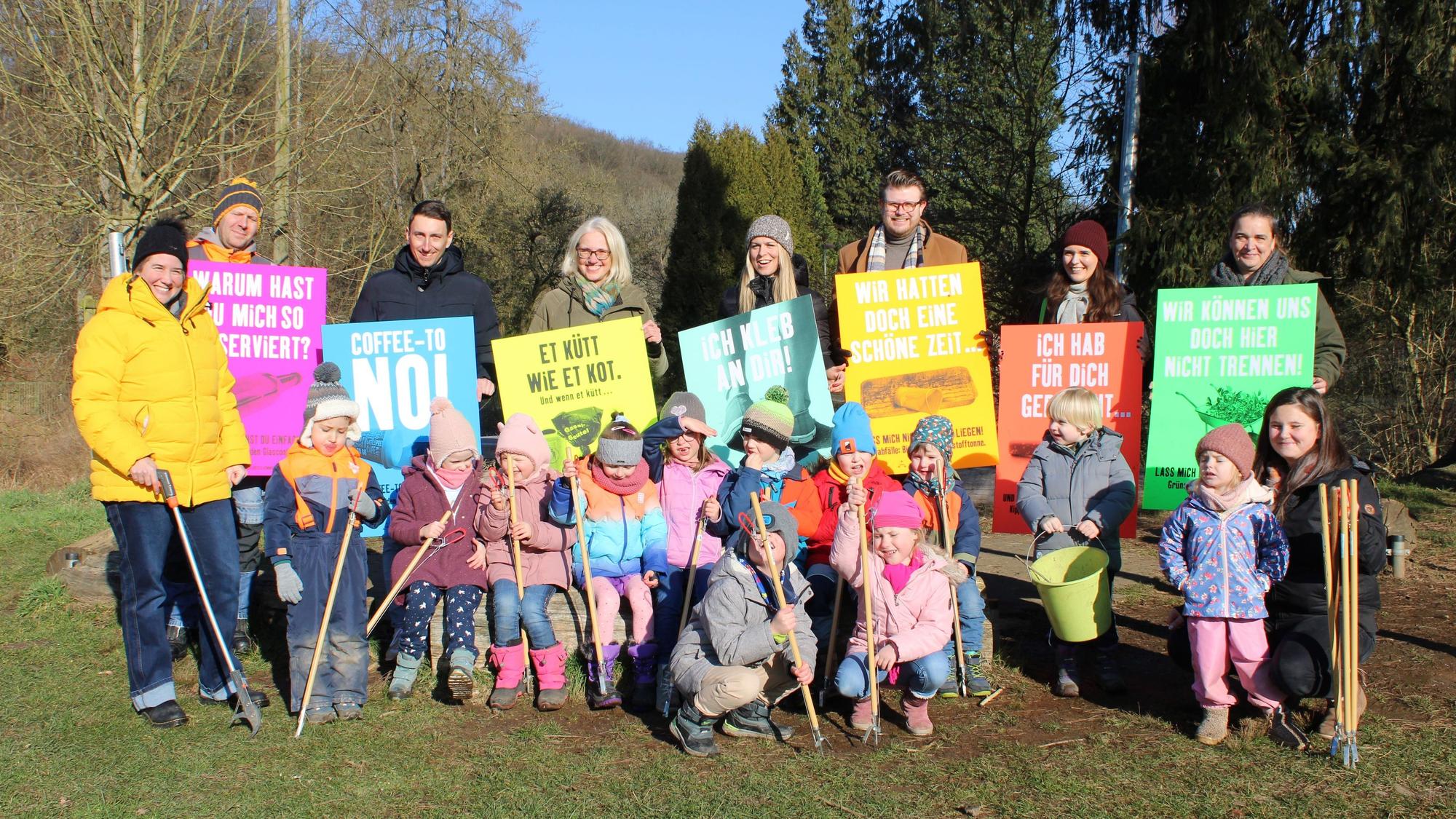 12 Kinder und 9 Erwachsene sind zu einem Gruppenfoto aufgestellt, die Kinder tragen Müllzangen in den Händen, die Erwachsenen halten Plakate, auf denen Beispielsweise „Coffee-to No!“ und „Et kütt wie et kot“ steht.