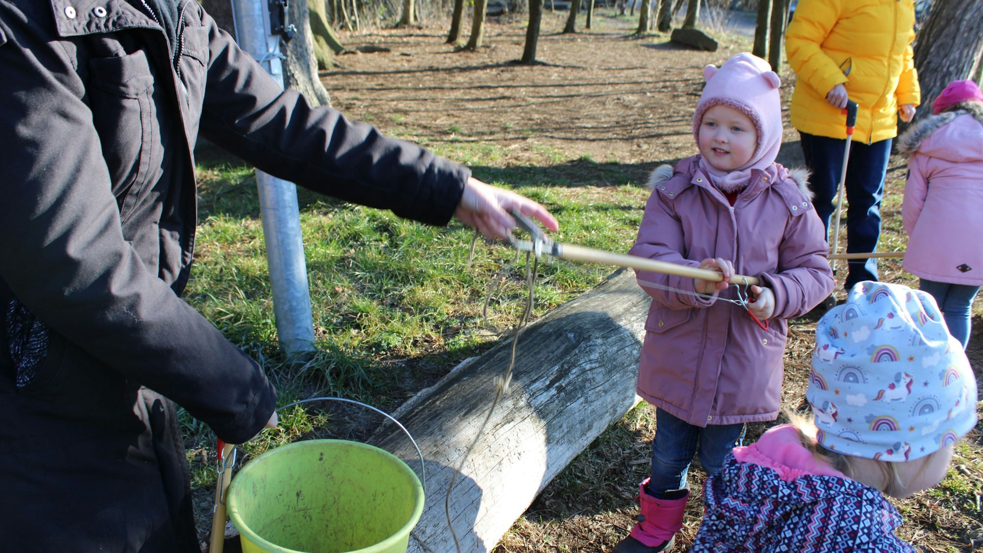 Ein Mädchen hebt mit seiner Müllzange eine lange Schnur hoch, eine Erzieherin hilft ihr, sie in den Plastikeimer zu legen.
