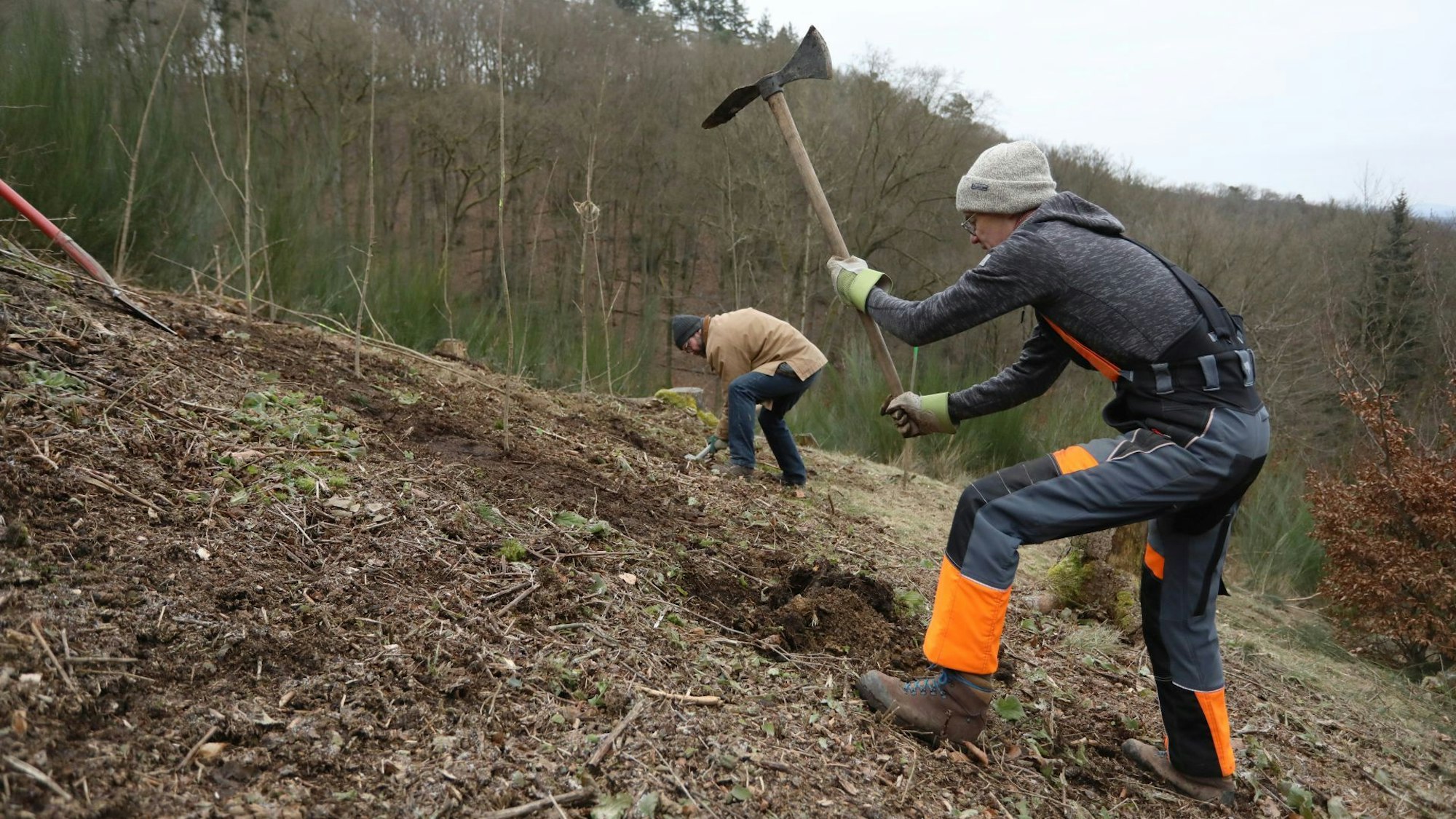 Zwei Männer arbeiten an einem Hang, einer hackt ein Loch in den Boden, der zweite im Hintergrund arbeitet mit einer Schaufel.