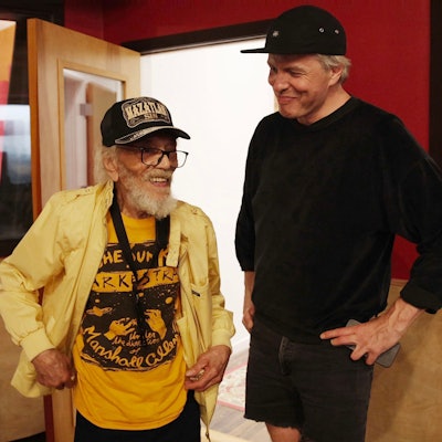 Jazz-Saxofonist Marshall Allen trägt ein gelbes offenes Hemd über einem dunkelgelben "Sun Ra Arkestra"-T-Shirt, rechts neben ihm steht Jan Lankisch, ganz in Schwarz, beide tragen Baseballkappen.