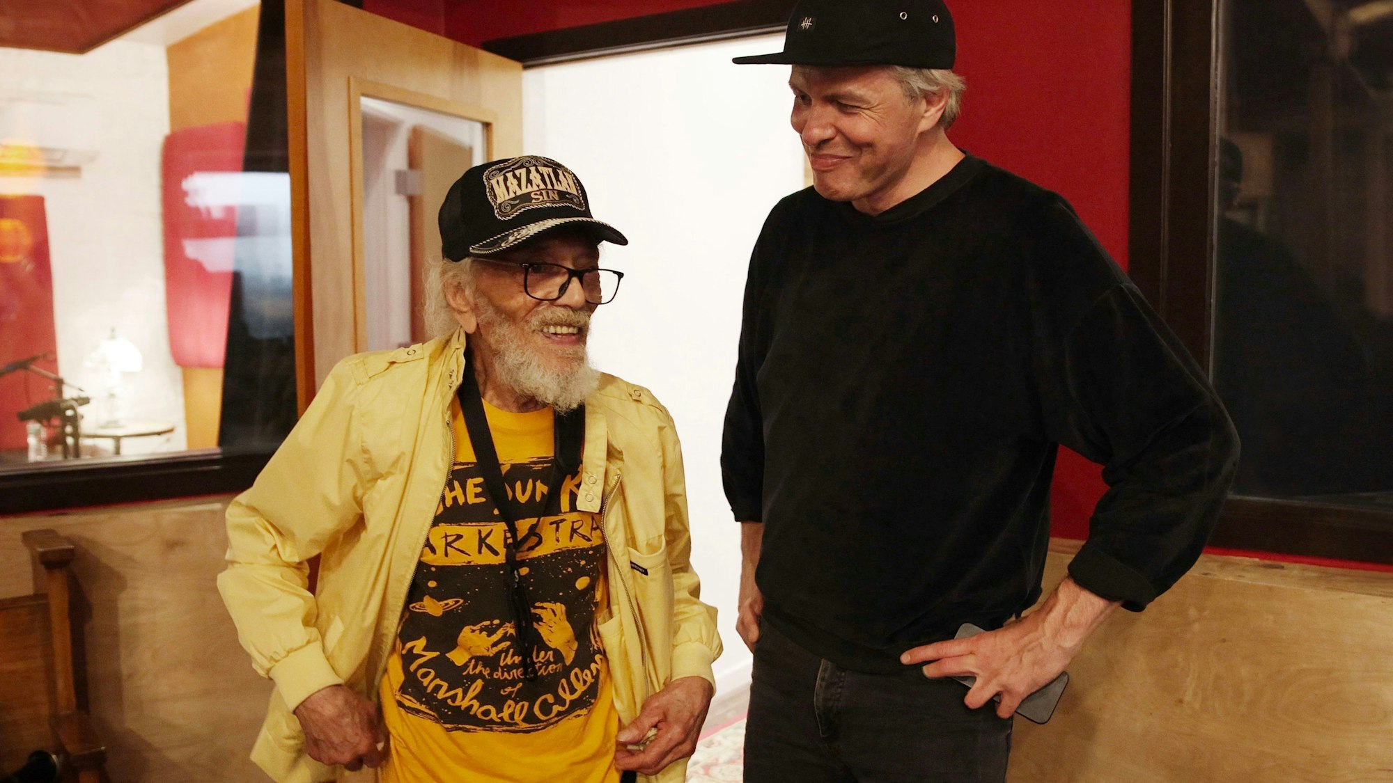 Jazz-Saxofonist Marshall Allen trägt ein gelbes offenes Hemd über einem dunkelgelben "Sun Ra Arkestra"-T-Shirt, rechts neben ihm steht Jan Lankisch, ganz in Schwarz, beide tragen Baseballkappen.
