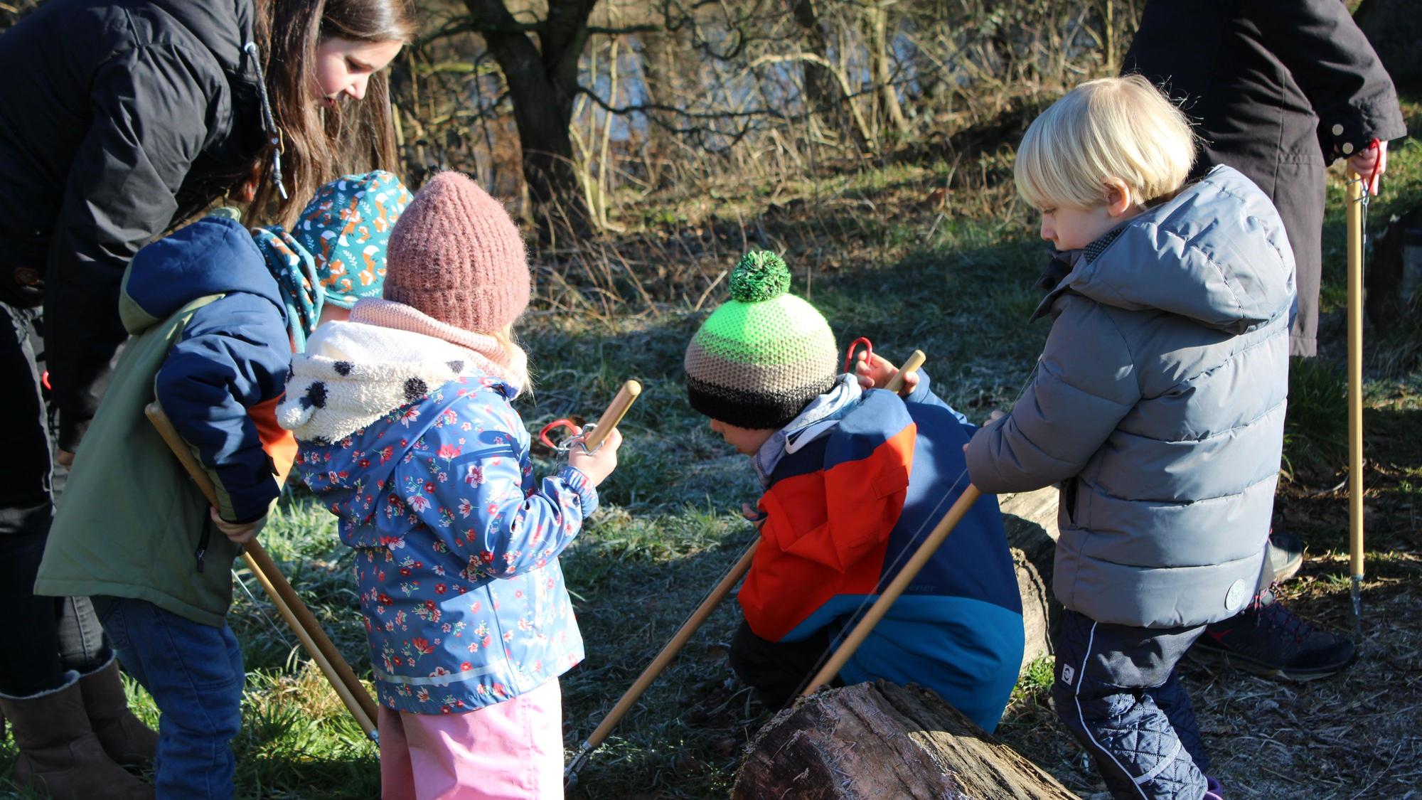 Vier Kinder suchen mit Müllzangen eine Wiese ab, eine Erzieherin steht neben ihnen.