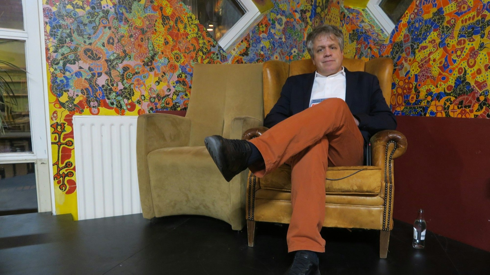 Ein grauhaariger Mann sitzt in einem Sessel vor einer bunten Wand.