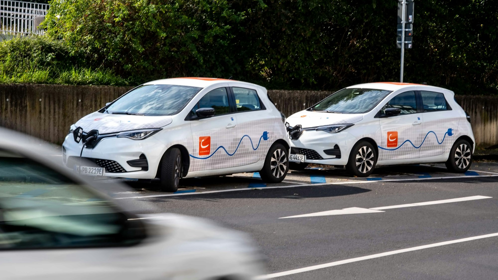 Zwei Fahrzeuge des Carsharing-Anbieters Cambio stehen am Straßenrand, ein Auto fährt vorbei.