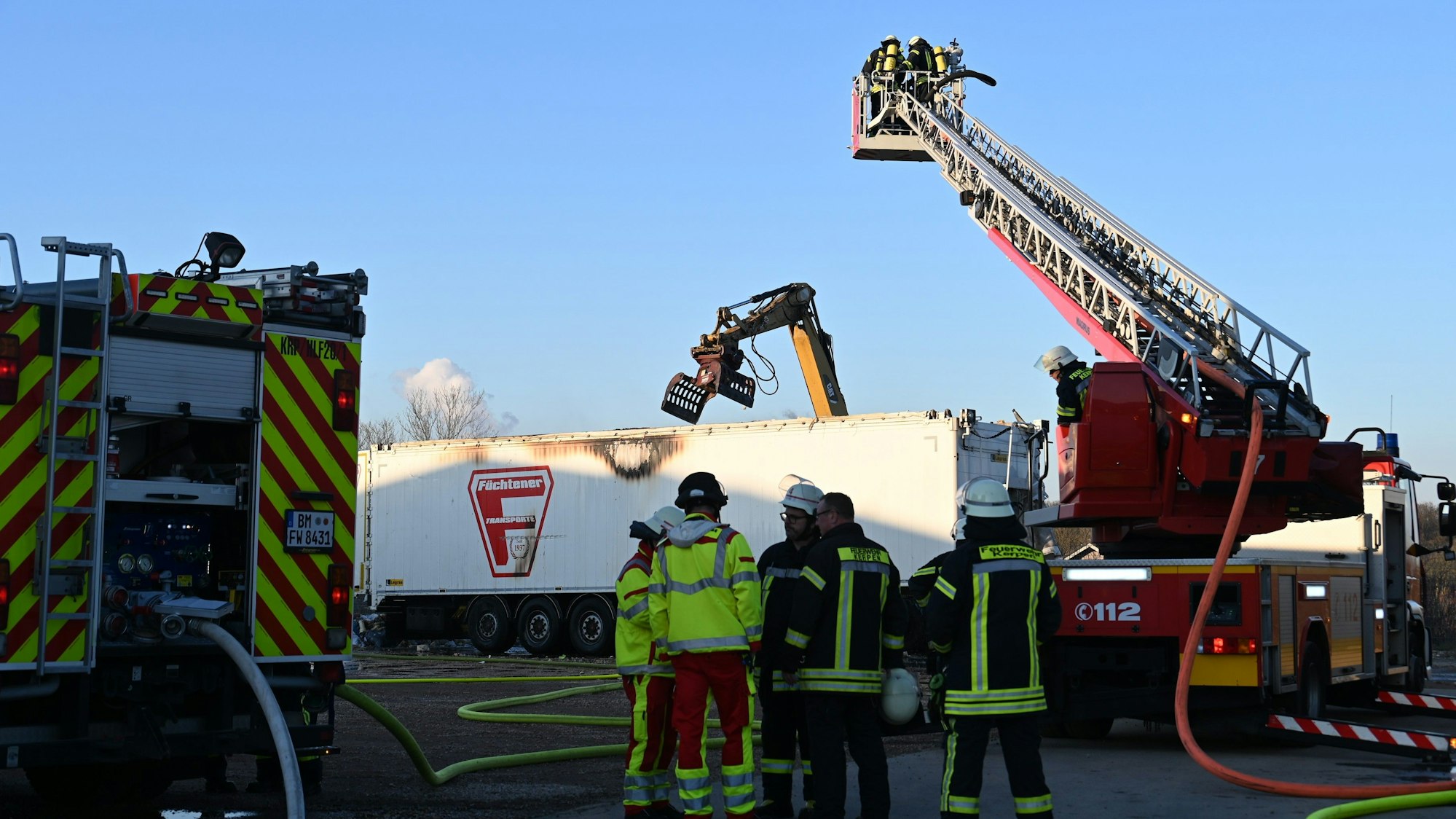 Feuerwehrleute stehen vor einem Lkw-Hänger. Neben ihnen steht ein Feuerwehrkran. Ein Bagger greift mit seiner Schaufel in den Hänger.