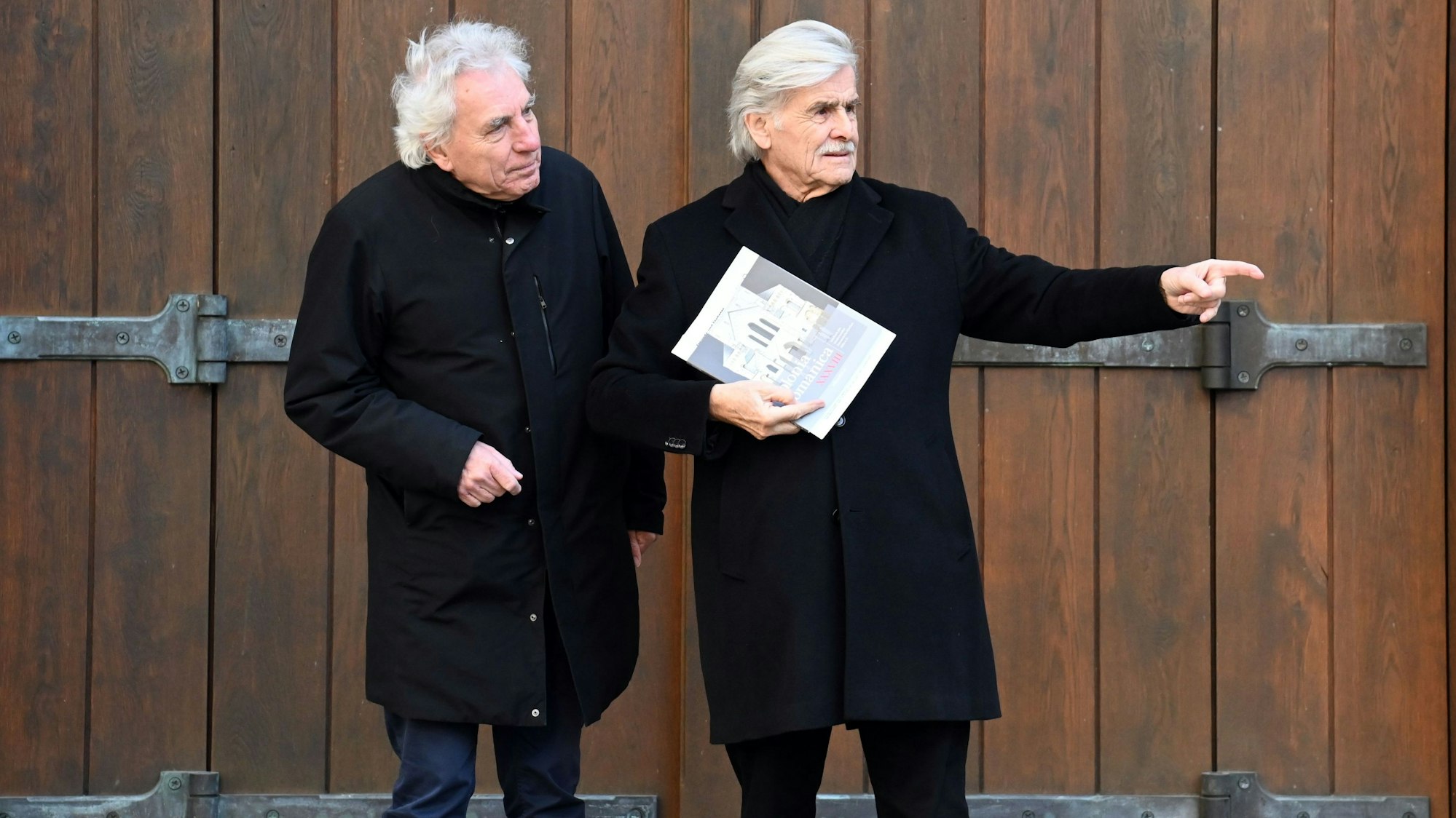 Jürgen Roters (links) und Helmut Haumann stehen vor einem geschlossenen Tor. Haumann zeigt in die Ferne.