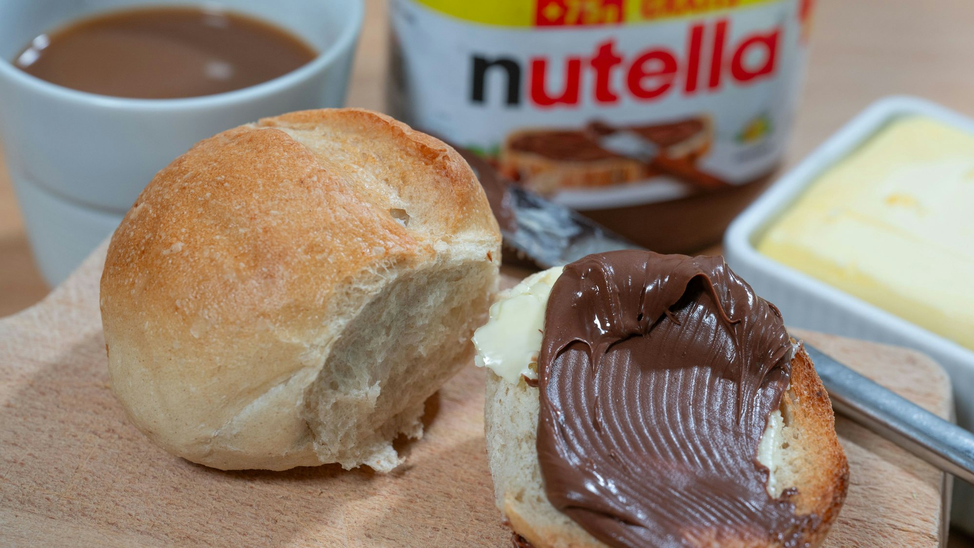Ein Brötchen mit Nutella liegt auf einem Frühstücksbrett.