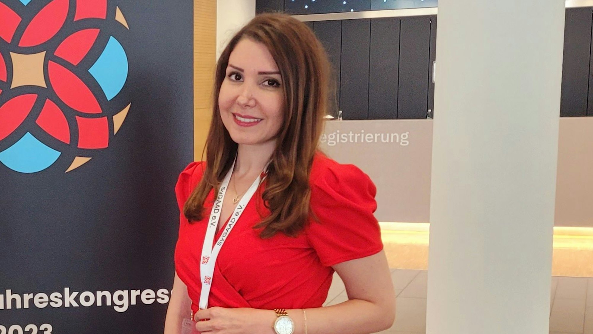 Nadia Abazid ist im Porträt zu sehen. Sie steht vor einem Kongress-Schild und hat eine rote Bluse an.