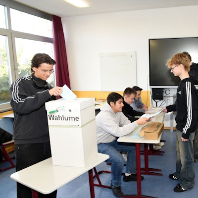 Mehrere Schüler stehen an einer Wahlurne in einem Klassenzimmer an, in die sie ihre Wahlzettel schmeißen.