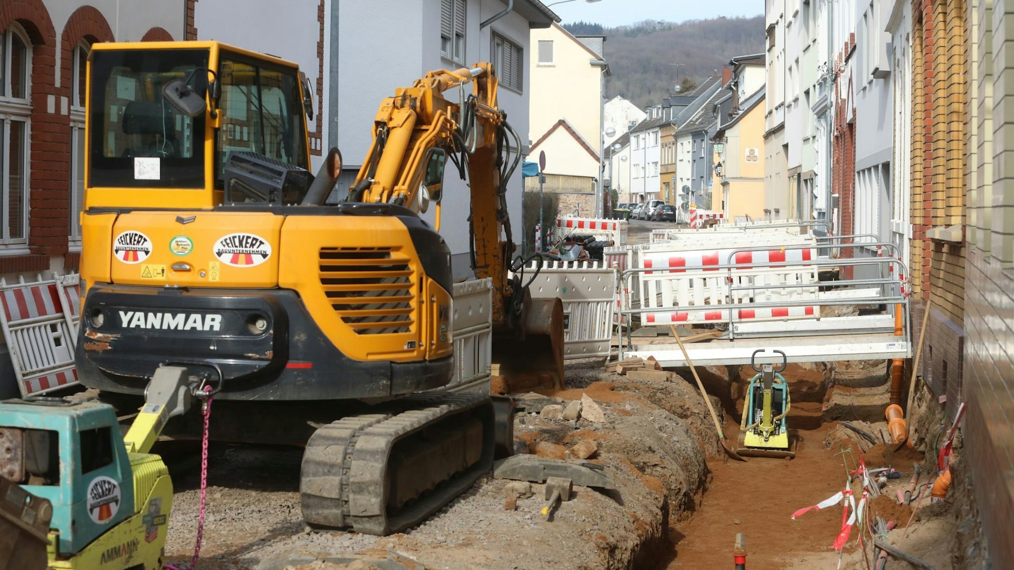 Ein gelber Bagger neben einer Baugrube in einer eng bebauten Straße.