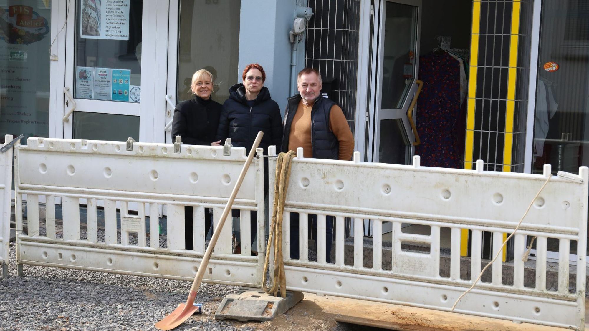 Drei Menschen hinter Speerbaken einer Baustelle, die nur wenig Platz zu den Ladenlokalen lassen.