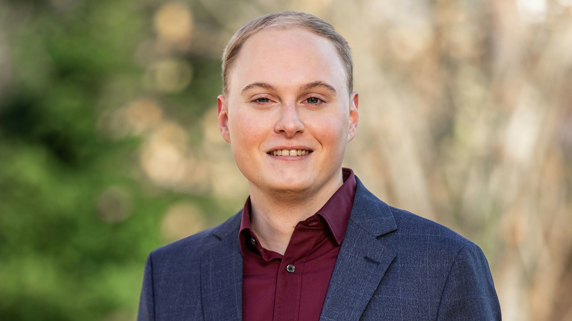 Das Foto zeigt einen jungen Mann mit blondem Haar. Er trägt ein blaues Sakko und ein bordeaux-rotes Hemd.
