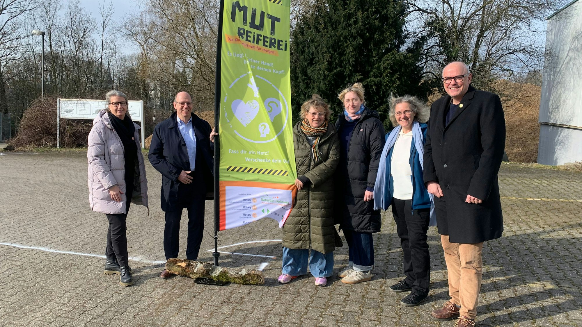 Mehrere Personen stehen nebeneinander an einer Fahne auf einem Parkplatz.