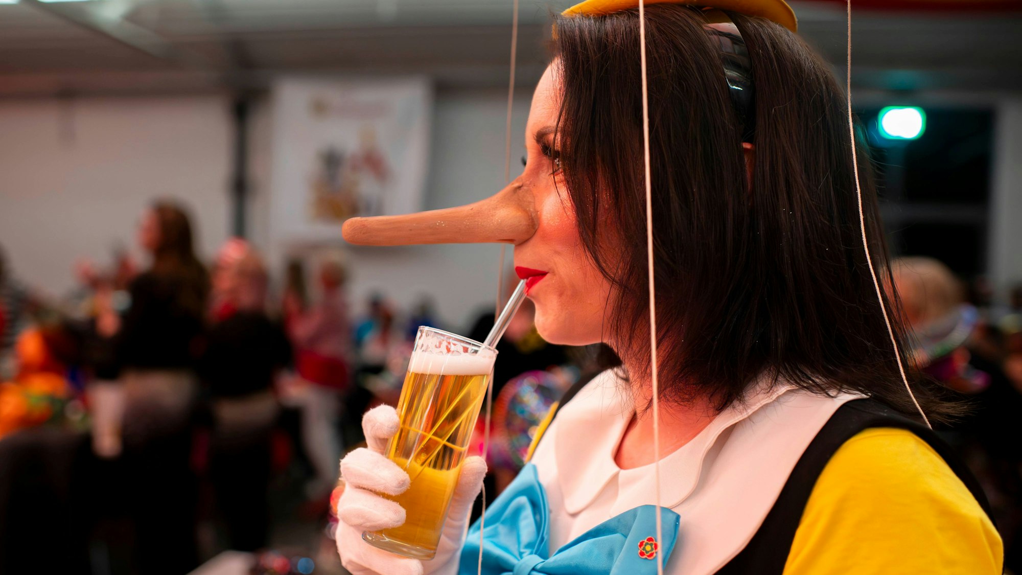 Das Bild zeigt die Obengenannte als Pinocchio verkleidet. Sie trinkt ein Bier mit dem Strohhalm.