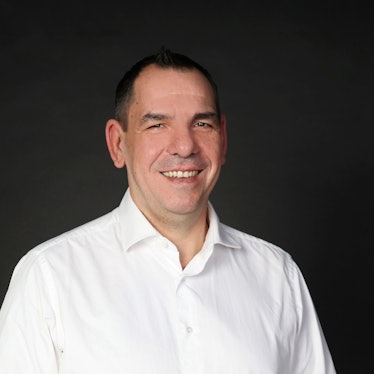 Das Bild zeigt Thomas Wiza. Er trägt ein weißes Hemd und steht vor einer schwarzen Wand.