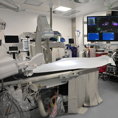 Dr. Erol Saygili, der Chefarzt der Kardiologie am Kreiskrankenhaus in Mechernich, trägt einen weißen Kittel und steht in einem Herzkatheter-Labor, das mit vielen technischen Geräten und einem großen Bildschirm ausgestattet ist.