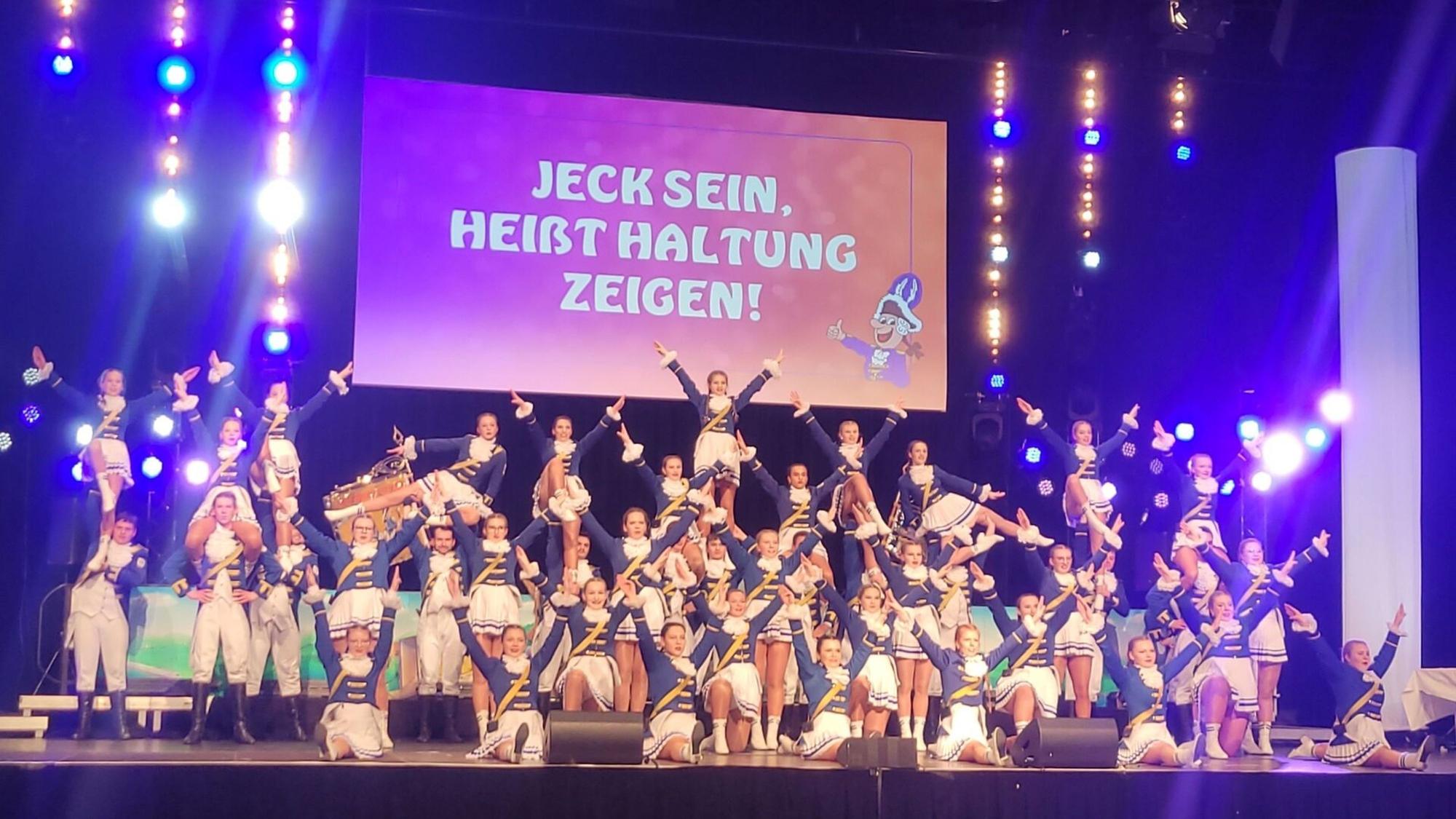 Eine Gardetanzgruppe auf der Bühne. Im Hintergrund hängt eine Leinwand mit der Aufschrift „Jeck sein heißt Haltung zeigen“.