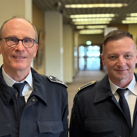 Auf dem Bild sind zwei Männer in Feuerwehruniform zu stehen, die in einem Flur des Frechener Rathauses stehen.