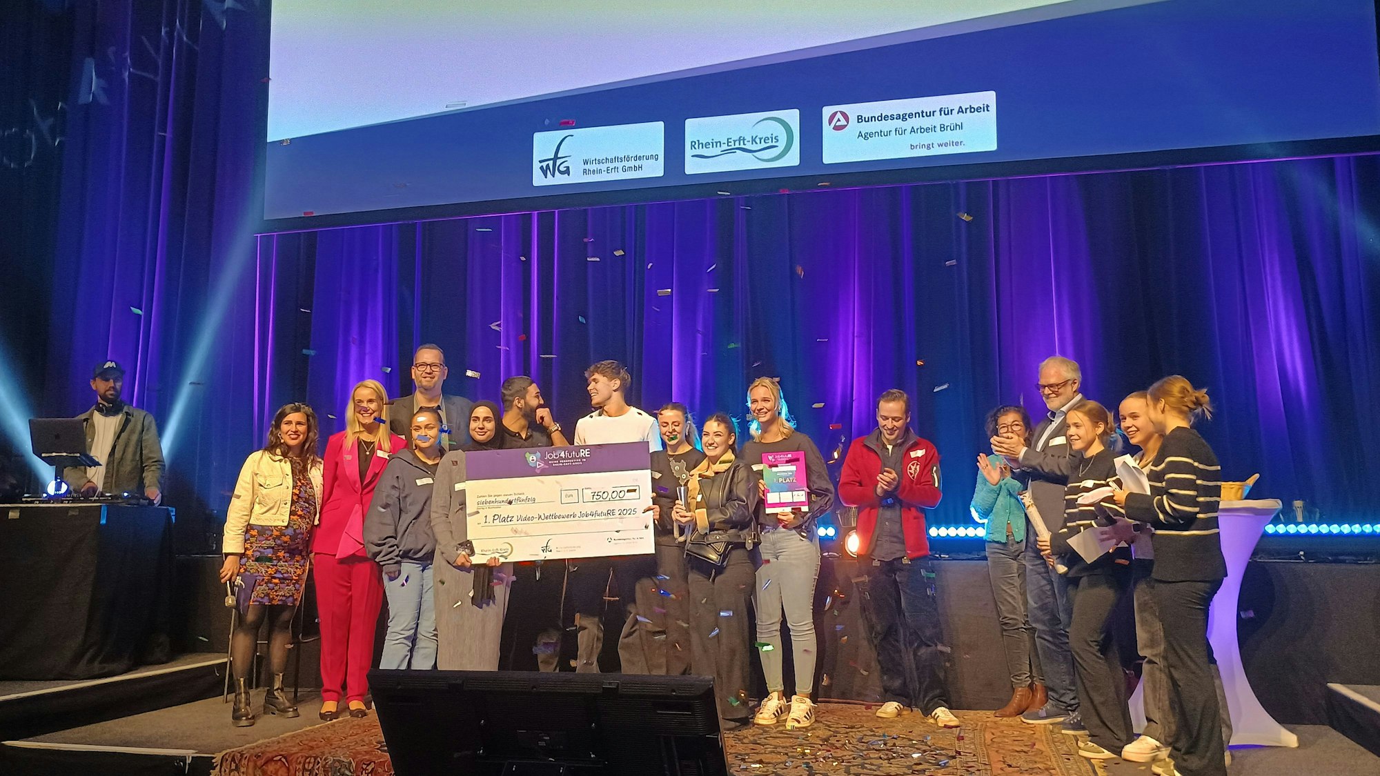 Die Schülerinnen und Schüler stehen mit der Jury auf der Bühne und halten ihren Scheck, während Konfetti herunterrieselt.
