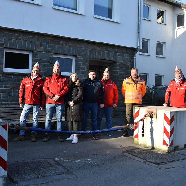 Mehrere Vereinsmitglieder eines Karnevalsvereins stehen auf der Straße neben einer Antiterrorsperre mit Sicherheitskette zwischen zwei Betonblöcken.
