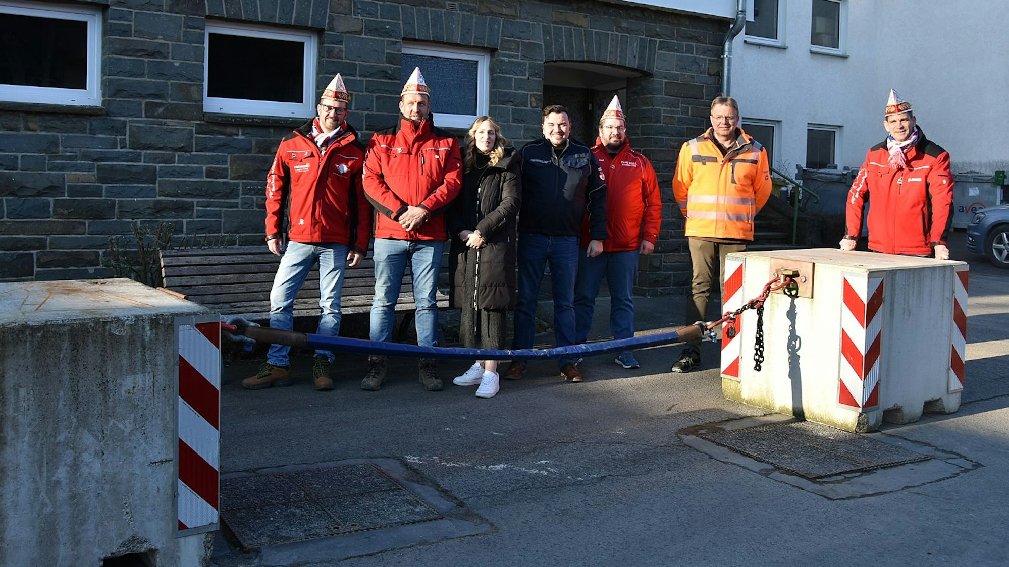 Mehrere Vereinsmitglieder eines Karnevalsvereins stehen auf der Straße neben einer Antiterrorsperre mit Sicherheitskette zwischen zwei Betonblöcken.