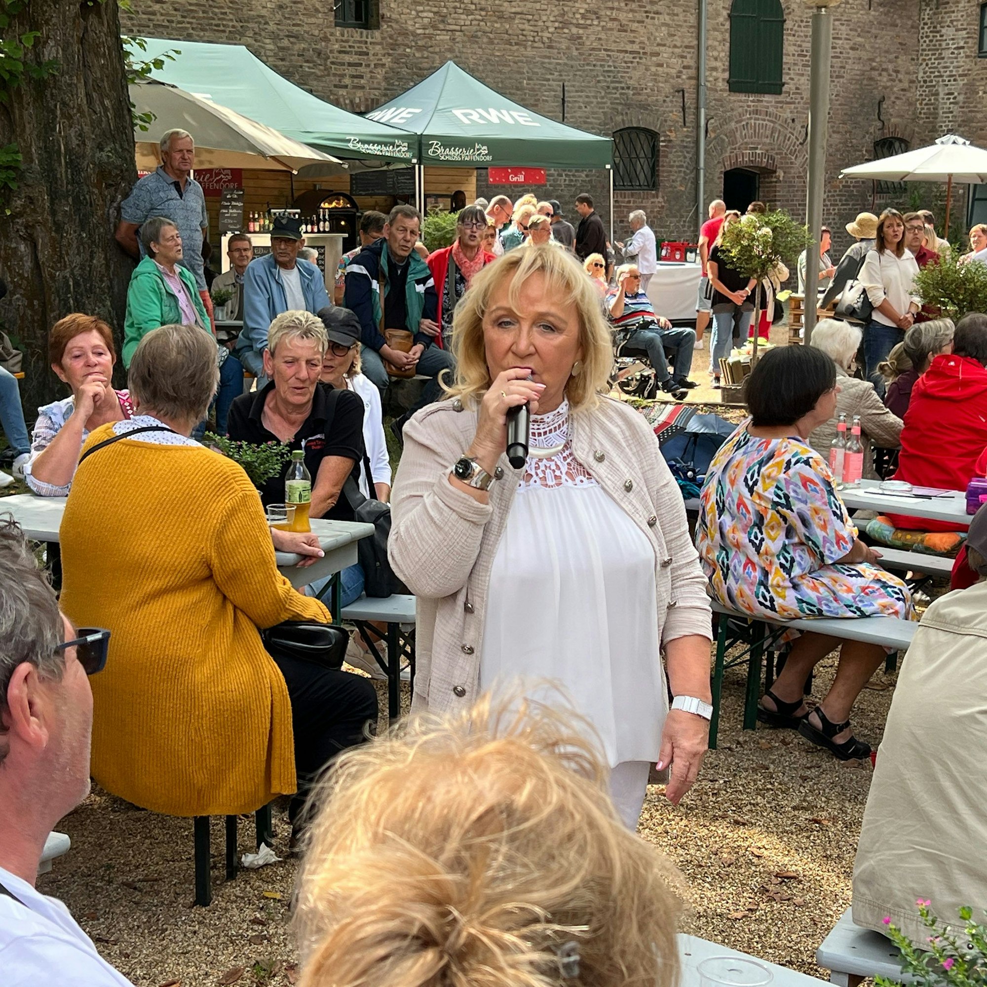 Eine Frau singt in einem Biergarten, in dem Menschen sitzen.