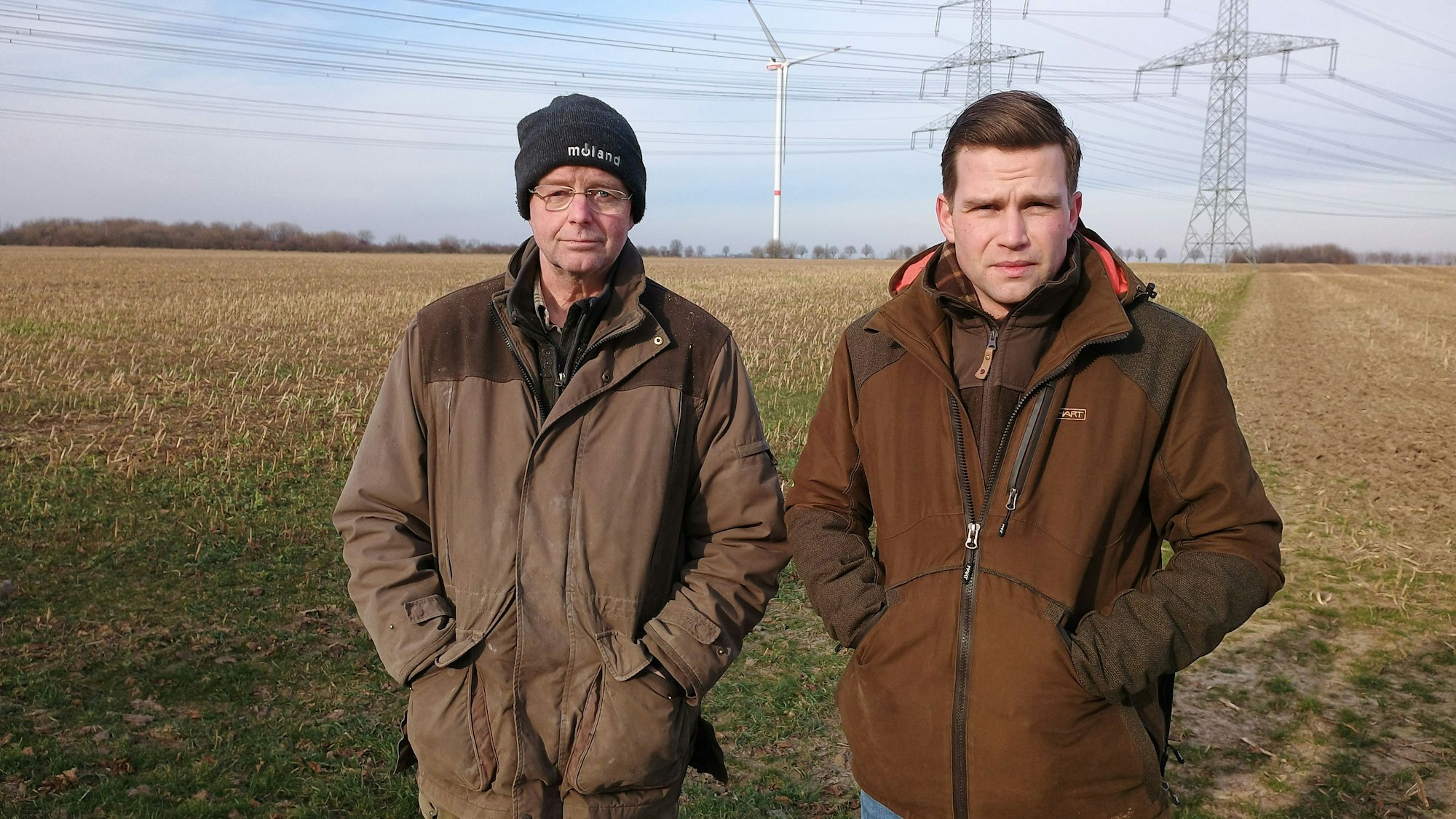 Die beiden stehen vor einem Feld und halten die Hände wegen der Kälte in ihre Jackentaschen. Im Hintergrund Stromleitungen und ein Windrad.