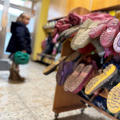 Ein Mädchen mit einem Fahrradhelm in der Hand steht im Eingangsbereich einer Kita, wo ein Gummistiefel-Ständer steht.