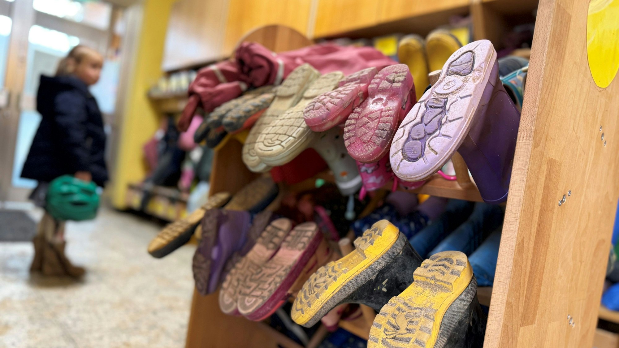 Ein Mädchen mit einem Fahrradhelm in der Hand steht im Eingangsbereich einer Kita, wo ein Gummistiefel-Ständer steht.