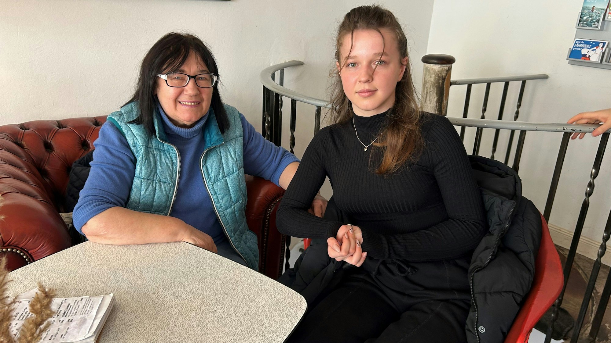 Nina (links) und Julia sitzen auf einer roten Couch und lächeln in die Kamera.