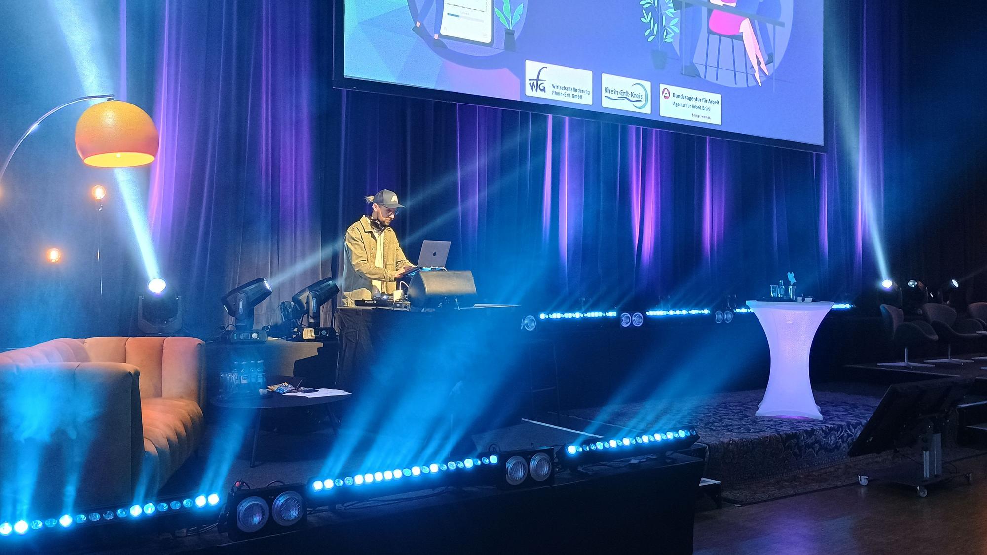 Das Bild zeigt den DJ an seinem Pult auf der Bühne, Scheinwerfer werfen sichtbare Lichtstreifen durch den Raum.
