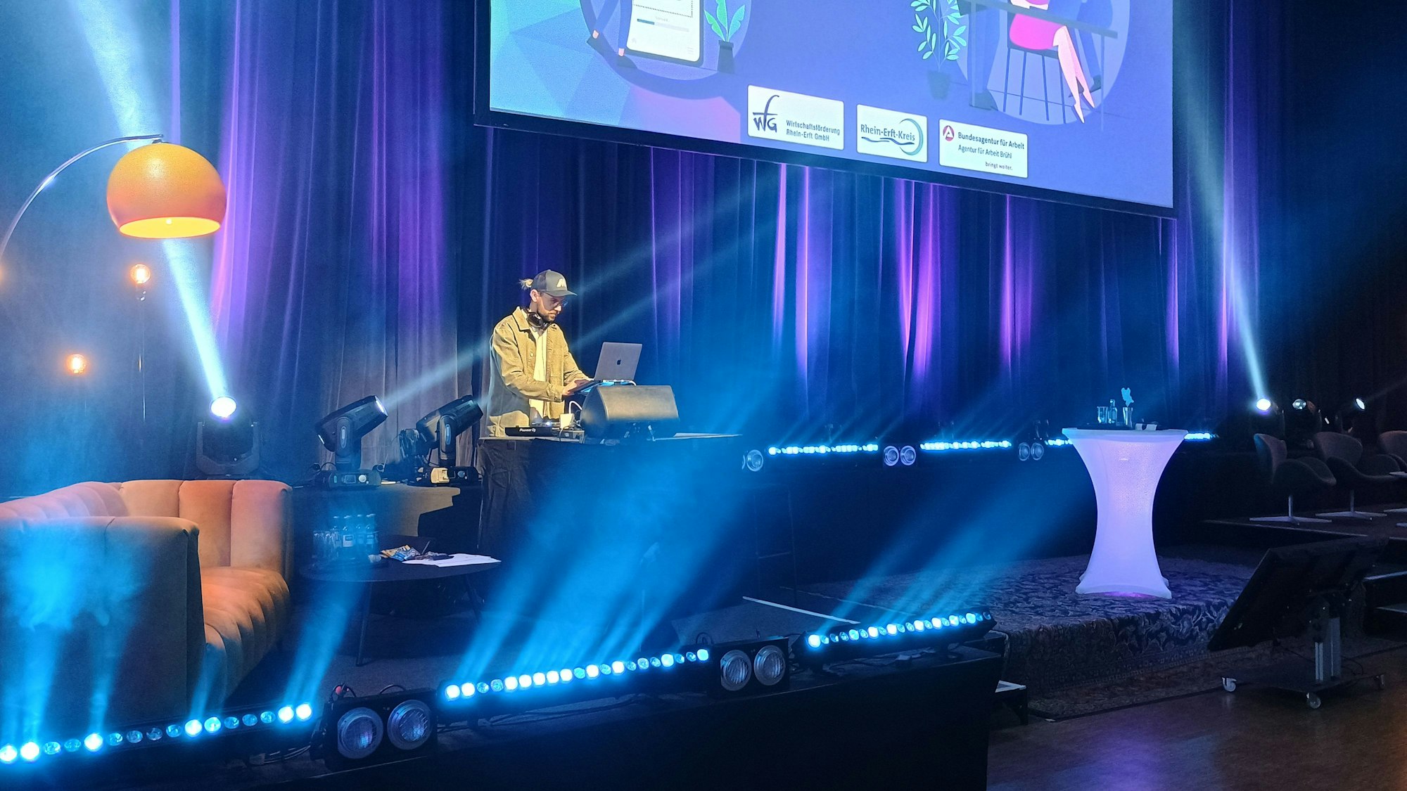 Das Bild zeigt den DJ an seinem Pult auf der Bühne, Scheinwerfer werfen sichtbare Lichtstreifen durch den Raum.