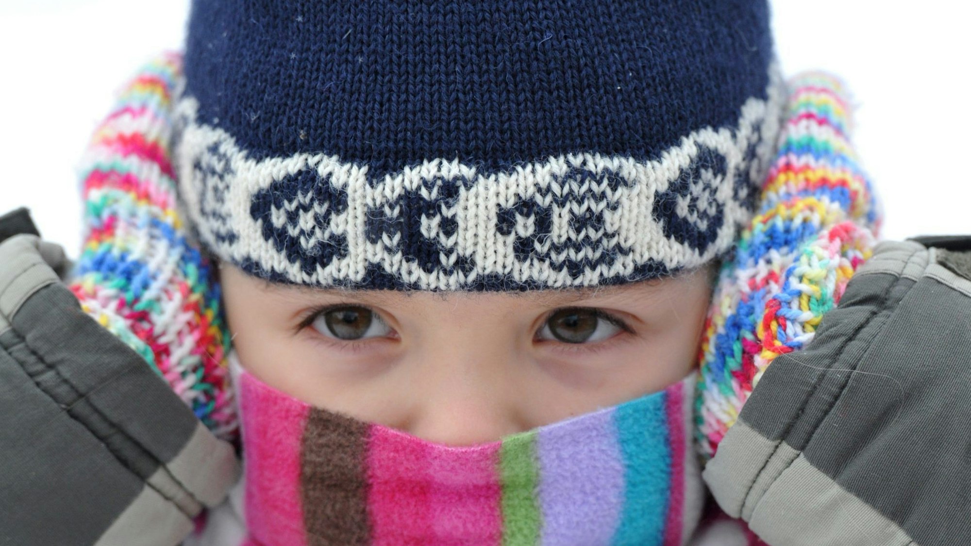 Ein kleines Mädchen hält sich bei klirrender Kälte schützend die Hände mit Handschuhen an ihre Ohren und trägt dabei eine Mütze sowie einen Schal.