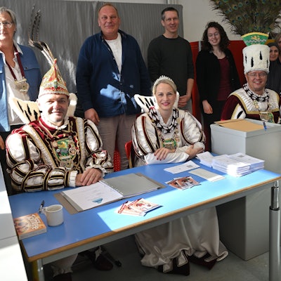 Das Dreigestirn sitzt am Tisch des Wahlvorstands in der Grundschule von Bergisch Gladbach-Gronau, dahinter stehen die ehrenamtliche Wahlhelfer.