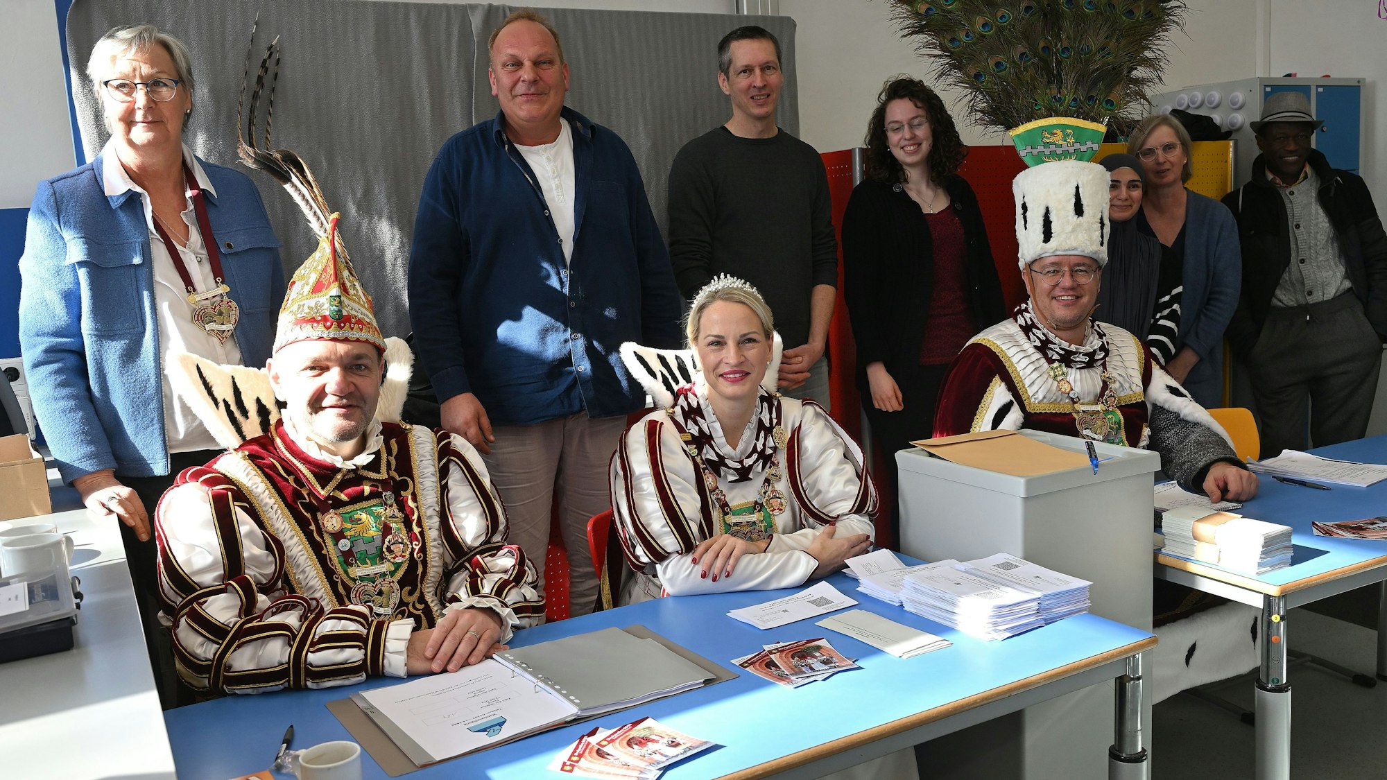 Das Dreigestirn sitzt am Tisch des Wahlvorstands in der Grundschule von Bergisch Gladbach-Gronau, dahinter stehen die ehrenamtliche Wahlhelfer.