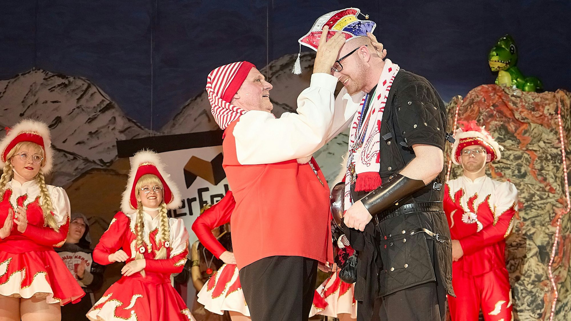 Auf der Bühne setzt ein Mann mit rot-weiß-geringelter Zipfelmütze dem Bürgermeister eine bunte Narrenkappe auf. Im Hintergrund stehen Mitglieder der Tanzgarde.