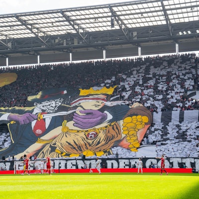 Ein Transparent, das Kölner Fans im Rhein-Energie-Stadion entfalteten, zeigt eine Comic-Figur, die einer Frau mit verbundenen Augen ein Messer an den Hals hält. Die Frau ist der Göttin Fortuna nachempfunden und schüttet Gold aus einem Füllhorn.