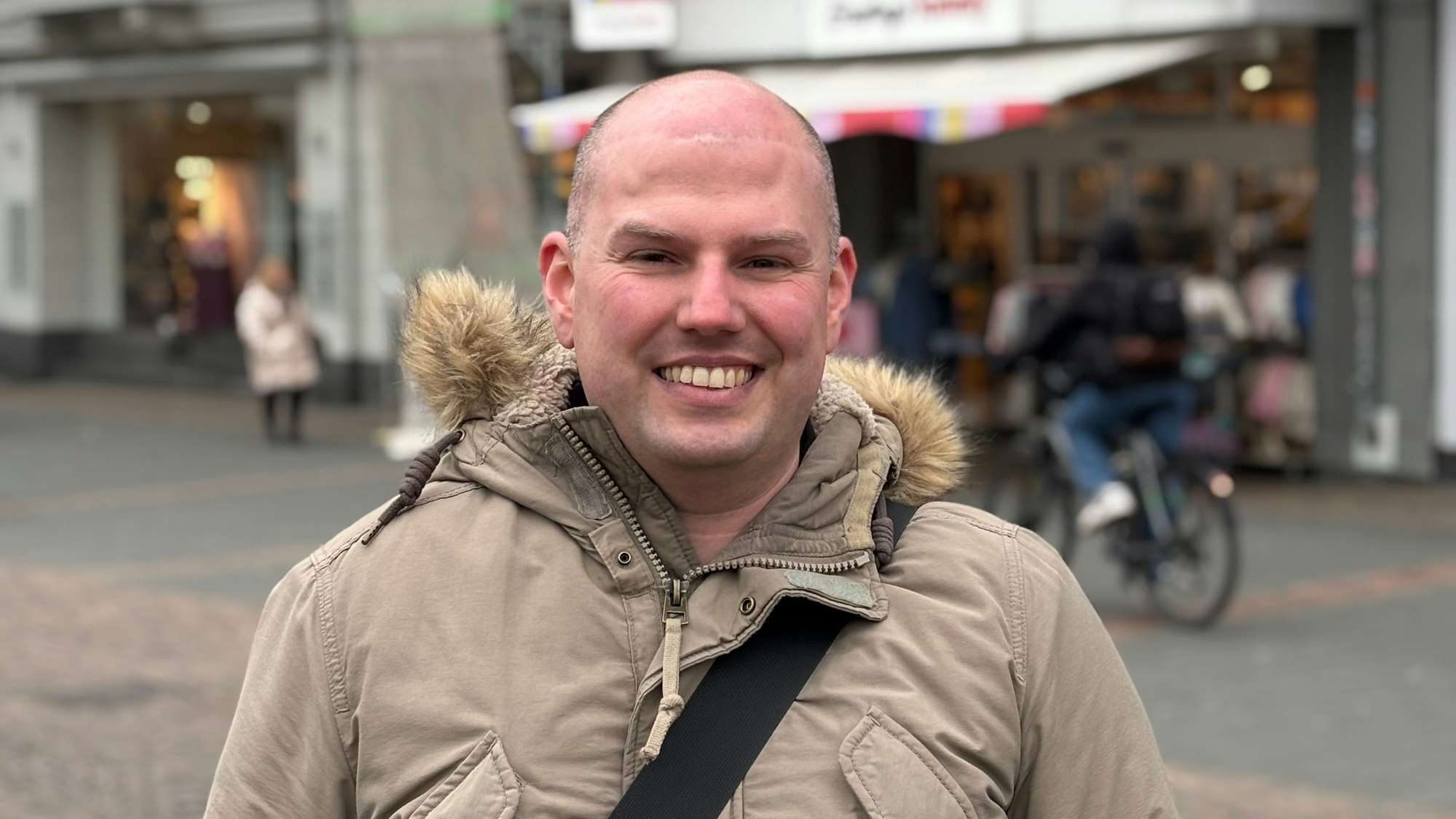 Ein Mann in einer Jacke steht in einer Fußgängerzone und zeigt seine Zähne beim Lächeln. Im Hintergrund befinden sich Passanten, die unscharf gestellt sind.