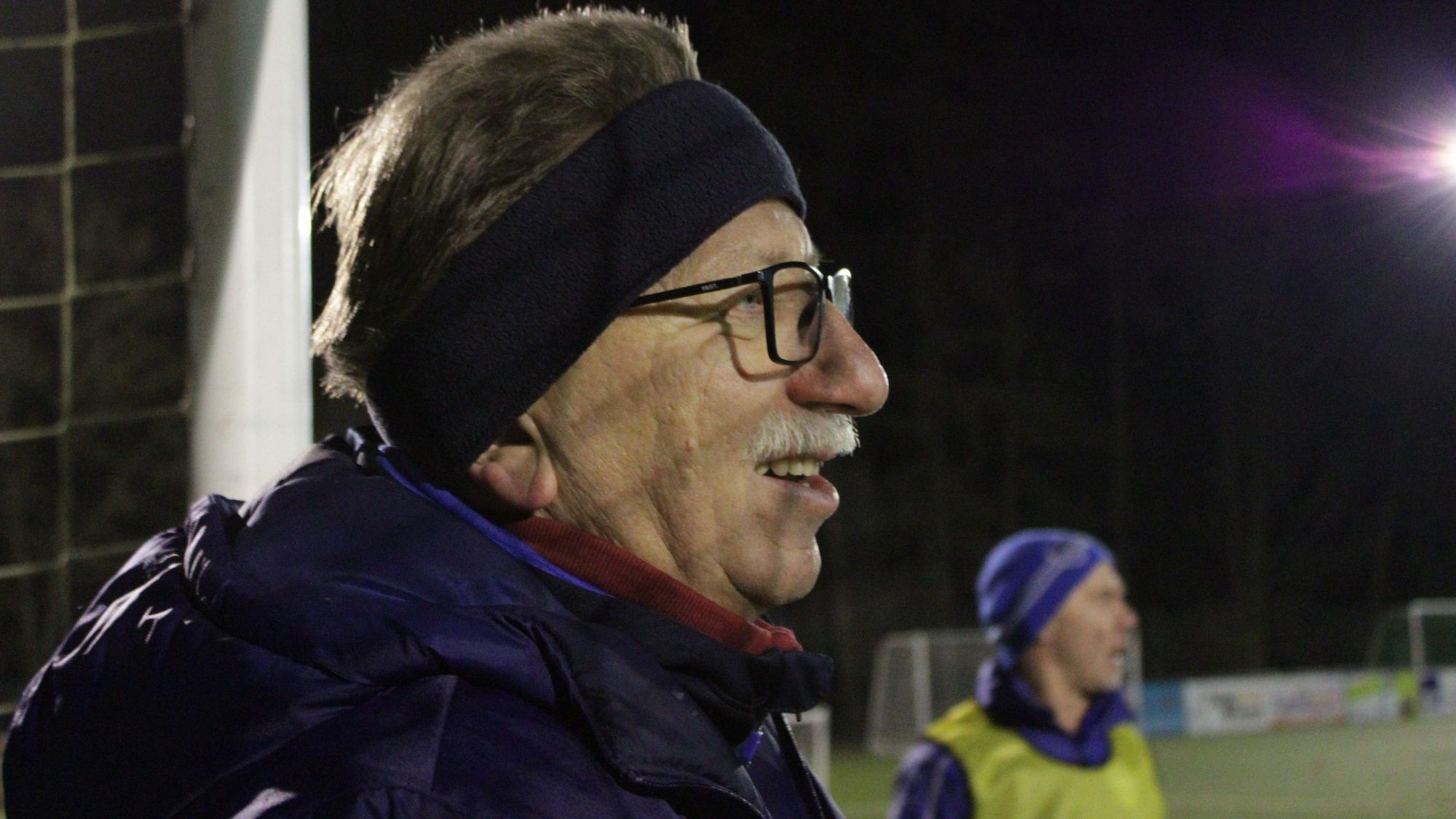 Ein älterer Mann mit Stirnband und Brille steht am Rand eines Fußballfeldes.