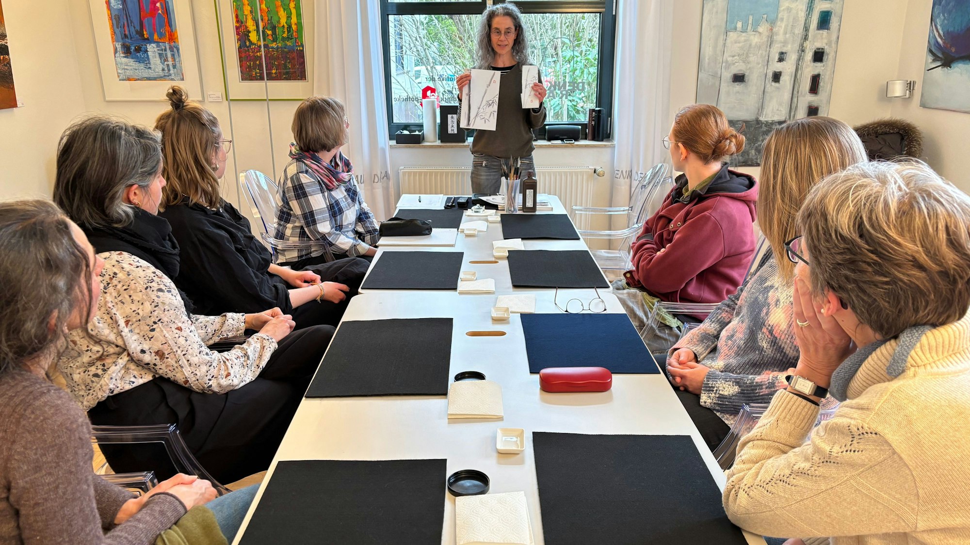 Eine Frau mit Brille steht vor sieben Frauen, die an einem Tisch sitzen und ihr zugewandt sind. Sie hält zwei Bilder in die Luft.