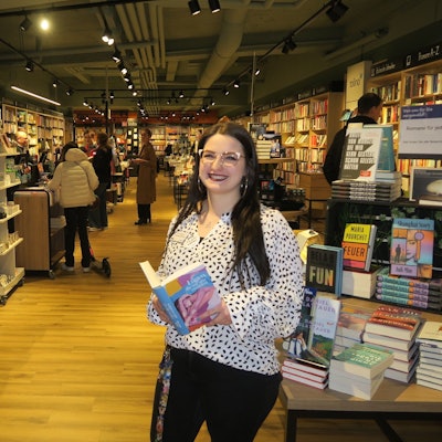 Eine junge Frau mit langem schwarzem Haar steht an einem Büchertisch mit einem aufgeschlagenen Buch in der Hand.