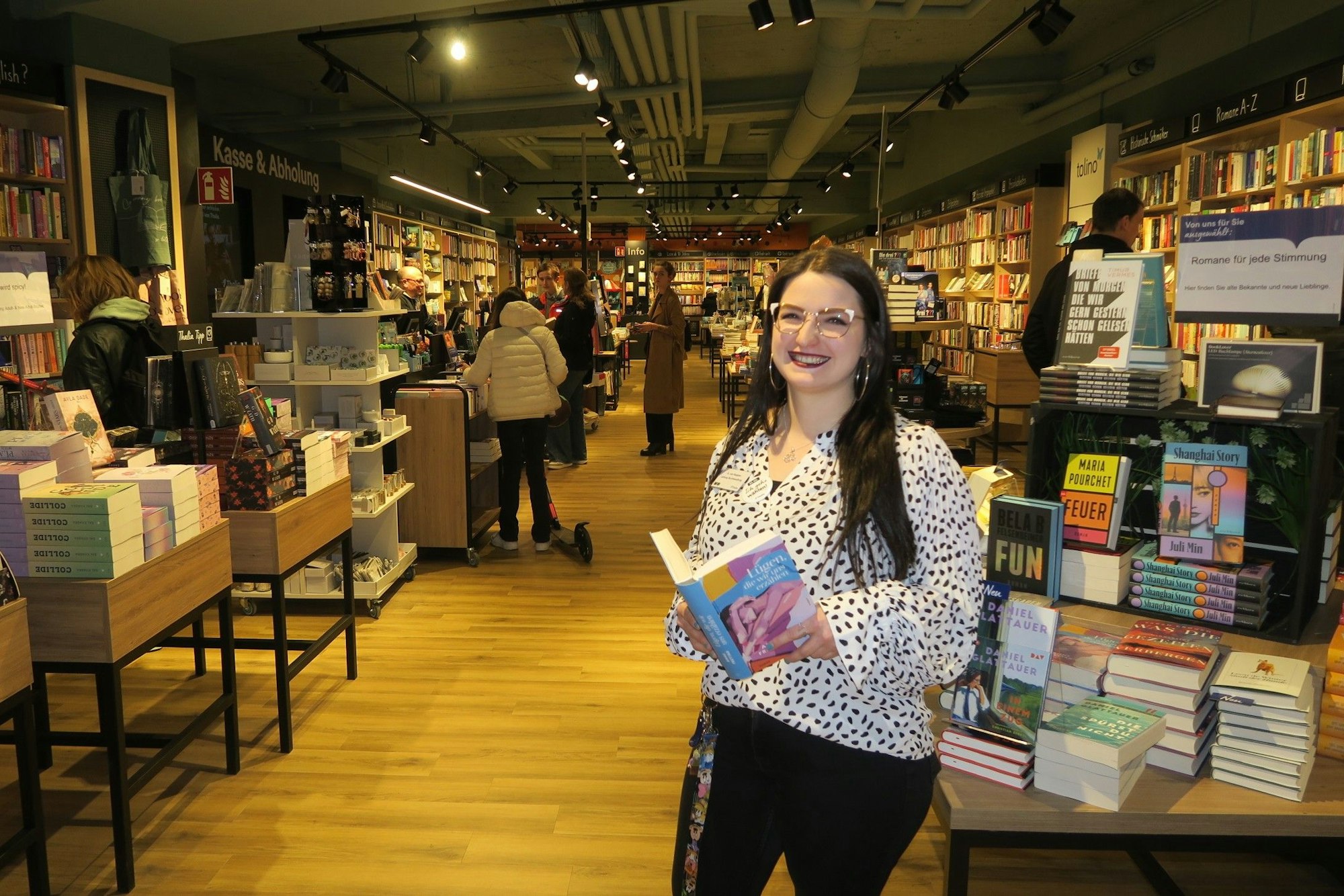 Eine junge Frau mit langem schwarzem Haar steht an einem Büchertisch mit einem aufgeschlagenen Buch in der Hand.