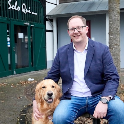 Sebastian Schubert sitzt auf einer Bank vor einem Baum. Mit einer Hand streichelt er einen Golden Retriever.