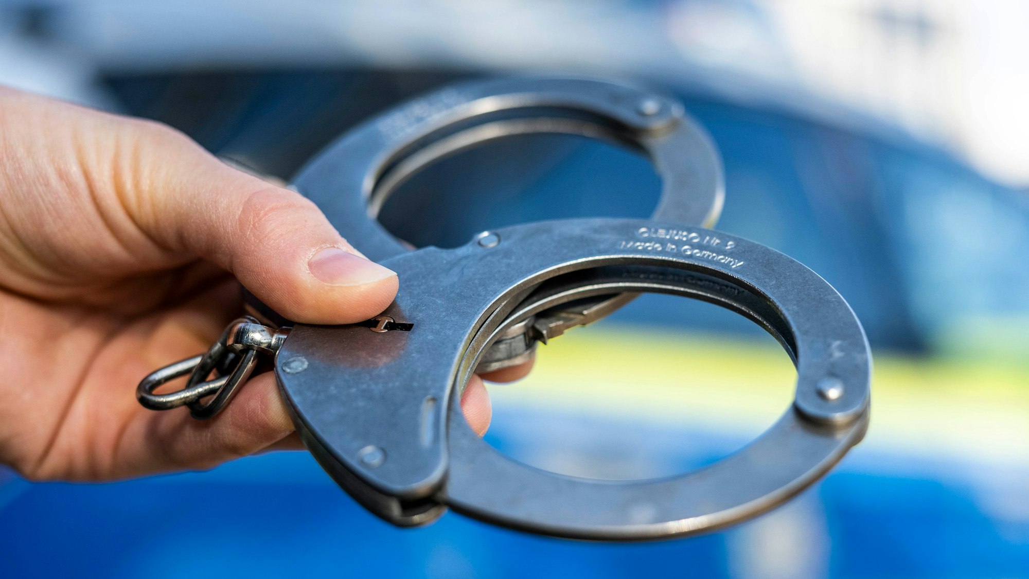 Eine Hand hält Handschellen in der Hand. Im Hintergrund verschwommen ein Polizeifahrzeug.