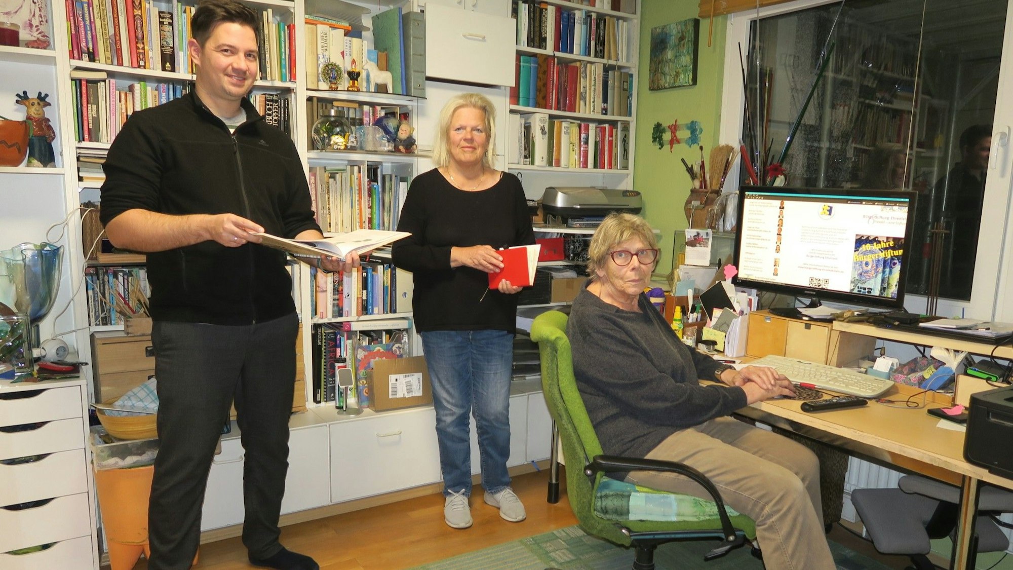Ein Mann und eine Frau stehen in einem Arbeitszimmer, eine ältere Frau sitzt an einem Schreibtisch mit großem Monitor.