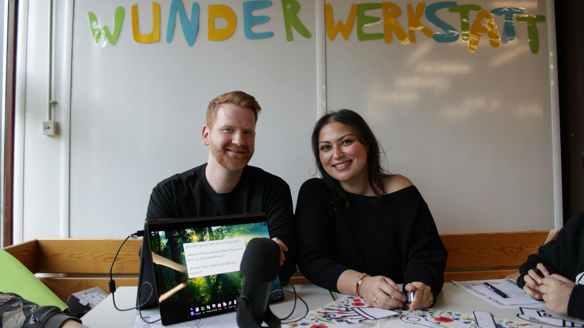 Ein Mann und eine Frau sitzen lächelnd an einem Tisch, hinter ihnen steht in bunten Buchstaben "Wunderwerkstatt" an der Wand, vor ihnen ist ein Mikrophon zu sehen.