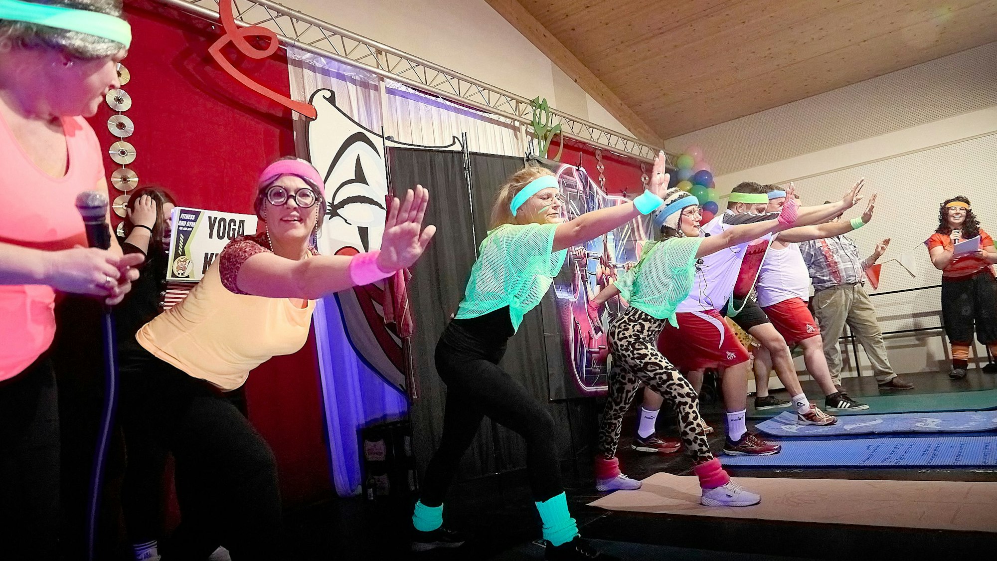 Frauen und Männer stehen in Sportkleidung auf einer Bühne und machen Übungen.