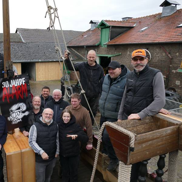 13 Männer und ein Mädchen in dunklen Klamotten stehen auf einem Holzschiff.