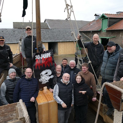 13 Männer und ein Mädchen in dunklen Klamotten stehen auf einem Holzschiff.