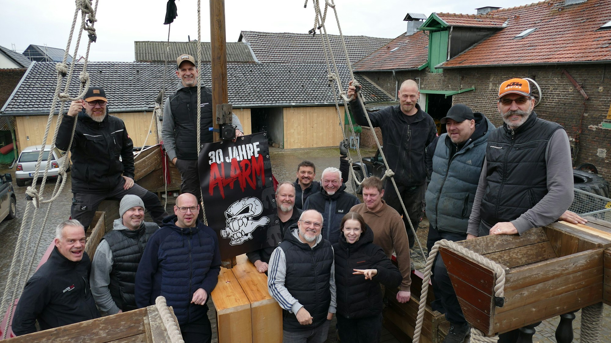 13 Männer und ein Mädchen in dunklen Klamotten stehen auf einem Holzschiff.